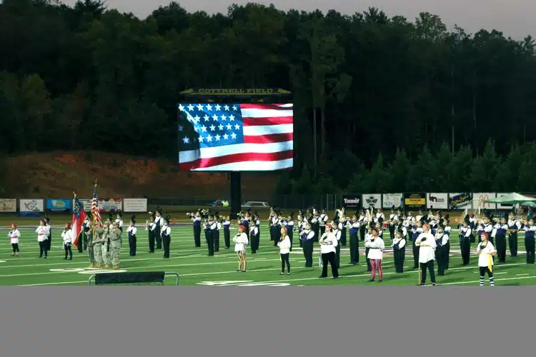 Marching band performing on field with an American flag scoreboard graphic